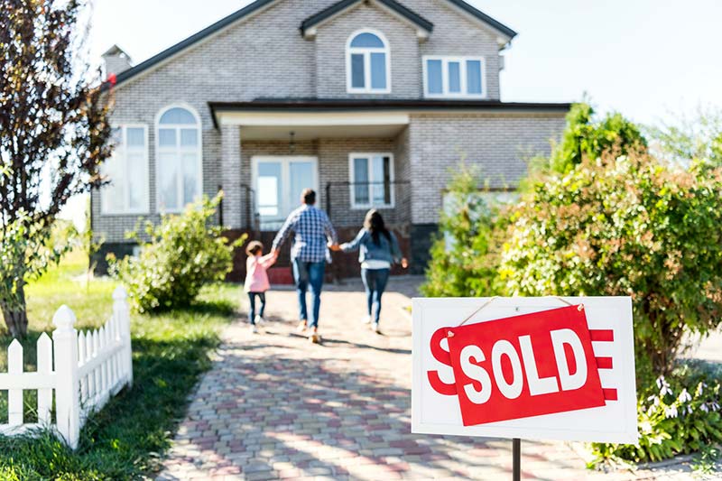 Home "for sale" sign under a "sold" sign and a family in the background going into the new house